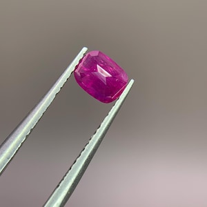 May include: A cushion-cut ruby gemstone, held by silver tweezers. The ruby exhibits a rich, pink-red colour with well-defined facets. The background is a soft, neutral grey.