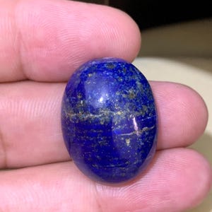 Puede incluir: Gema ovalada de lapislázuli pulido. La piedra azul oscuro presenta inclusiones doradas y una superficie lisa y brillante. La gema se sostiene en una mano, destacando su belleza natural y variaciones de color. Vista de cerca de la textura.