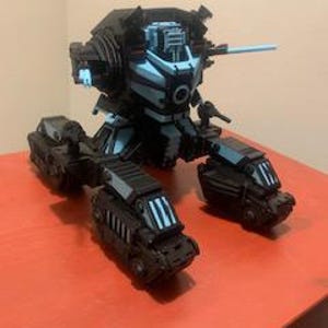 May include: A black and light blue robotic action figure featuring a central torso with a mounted cannon, articulated limbs, and tank-like treaded feet. The mechanical design includes geometric paneling, a prominent circular chest detail, and angular armor plating throughout the structure.