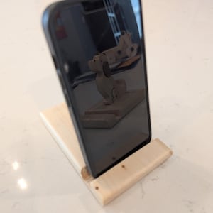 Puede incluir: Un soporte de tel&eacute;fono de madera clara, inclinado, sosteniendo un smartphone negro. Se aprecia un peque&ntilde;o perro de madera reflejado en la pantalla del tel&eacute;fono. El soporte es de madera de color claro.