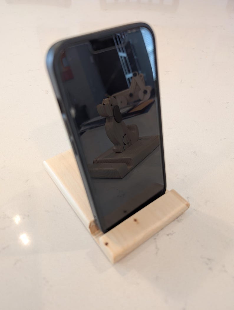 Puede incluir: Un soporte de tel&eacute;fono de madera clara, inclinado, sosteniendo un smartphone negro. Se aprecia un peque&ntilde;o perro de madera reflejado en la pantalla del tel&eacute;fono. El soporte es de madera de color claro.