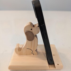 Puede incluir: Un soporte de tel&eacute;fono de madera con un dise&ntilde;o de perro tallado. El soporte est&aacute; hecho de madera de color claro y sostiene un tel&eacute;fono inteligente negro. El perro tiene acentos de oreja marrones y detalles de contorno negros.