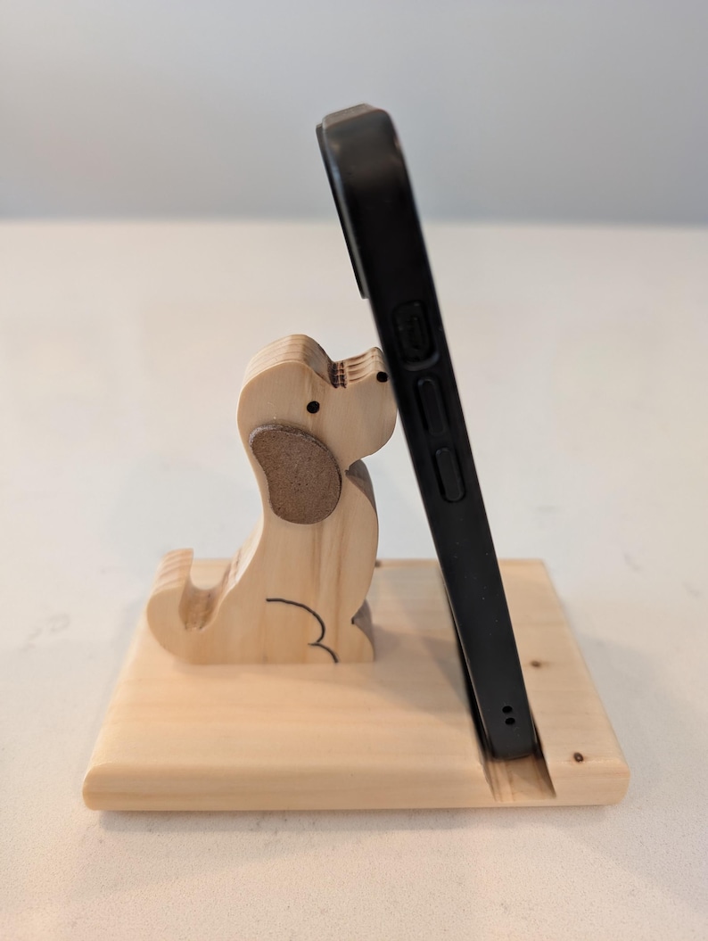 Puede incluir: Un soporte de tel&eacute;fono de madera con un dise&ntilde;o de perro tallado. El soporte est&aacute; hecho de madera de color claro y sostiene un tel&eacute;fono inteligente negro. El perro tiene acentos de oreja marrones y detalles de contorno negros.