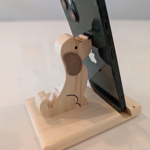 Puede incluir: Un soporte de tel&eacute;fono de madera con forma de perro, sosteniendo un smartphone negro. El perro tiene un cuerpo marr&oacute;n claro, una oreja marr&oacute;n y detalles negros. El soporte es de madera clara y tiene una base rectangular.