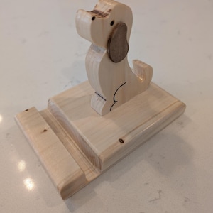 Puede incluir: Un soporte para tel&eacute;fono con forma de perro de madera. El soporte est&aacute; hecho de madera de color claro y presenta una silueta de perro con una oreja marr&oacute;n. La base tiene una ranura para sostener un tel&eacute;fono. El soporte mide aproximadamente 15 cm de largo y 10 cm de ancho.