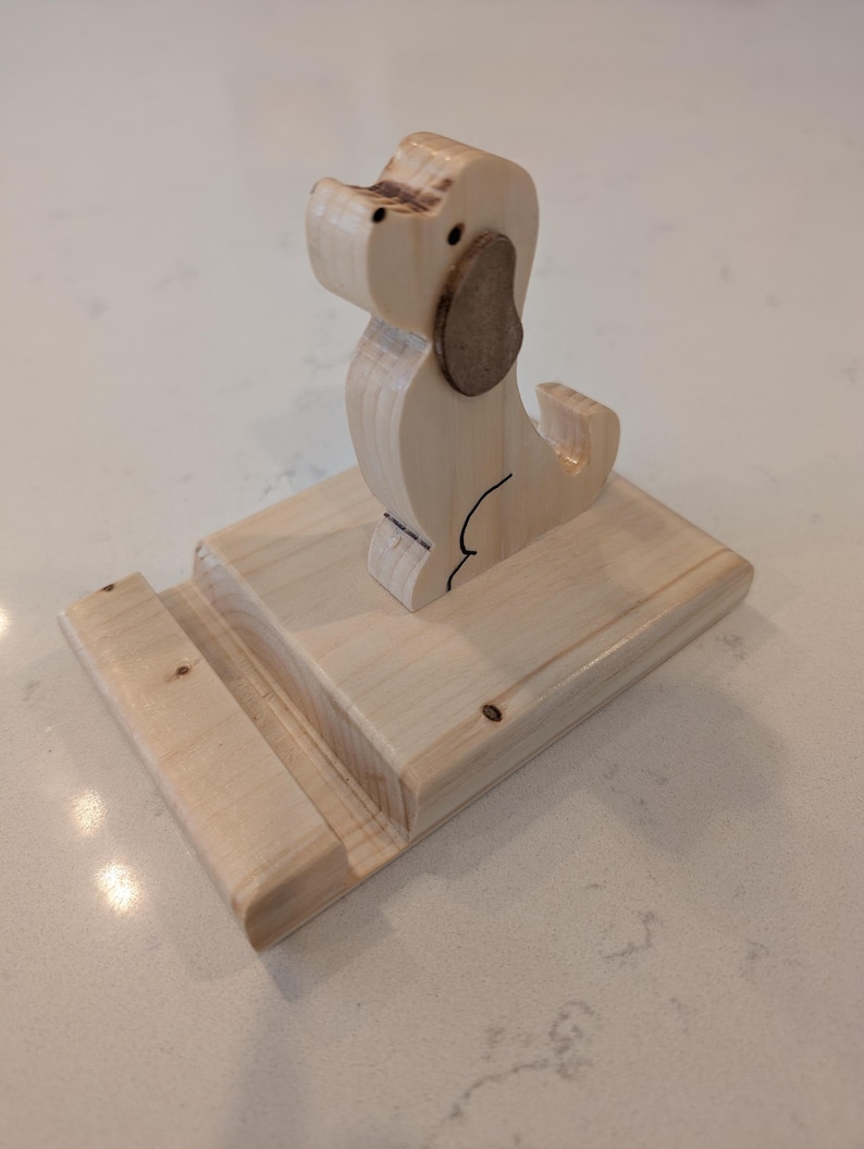 Puede incluir: Un soporte para tel&eacute;fono con forma de perro de madera. El soporte est&aacute; hecho de madera de color claro y presenta una silueta de perro con una oreja marr&oacute;n. La base tiene una ranura para sostener un tel&eacute;fono. El soporte mide aproximadamente 15 cm de largo y 10 cm de ancho.
