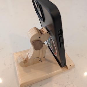 Puede incluir: Un soporte de tel&eacute;fono de madera con forma de perro, sosteniendo un tel&eacute;fono negro. El soporte est&aacute; hecho de madera clara con un detalle de oreja marr&oacute;n. El tel&eacute;fono tiene una pegatina colorida.