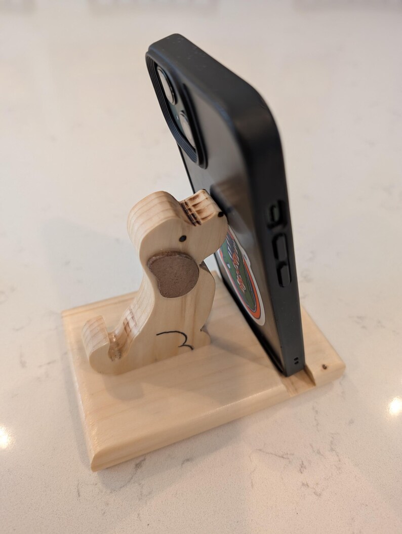 Puede incluir: Un soporte de tel&eacute;fono de madera con forma de perro, sosteniendo un tel&eacute;fono negro. El soporte est&aacute; hecho de madera clara con un detalle de oreja marr&oacute;n. El tel&eacute;fono tiene una pegatina colorida.