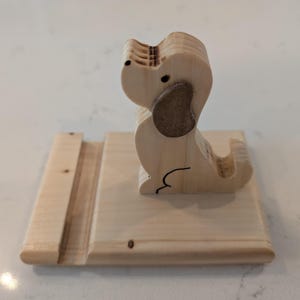 Puede incluir: Un soporte para tel&eacute;fono de madera con forma de perro. El perro tiene una oreja marr&oacute;n claro y un contorno negro. El soporte est&aacute; hecho de madera clara y tiene una ranura para sostener un tel&eacute;fono. El perro est&aacute; sentado sobre una base rectangular.