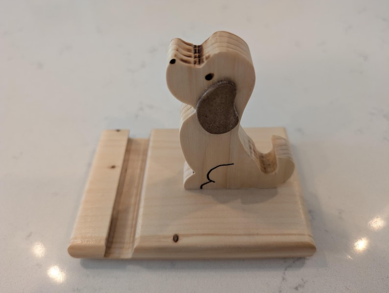 Puede incluir: Un soporte para tel&eacute;fono de madera con forma de perro. El perro tiene una oreja marr&oacute;n claro y un contorno negro. El soporte est&aacute; hecho de madera clara y tiene una ranura para sostener un tel&eacute;fono. El perro est&aacute; sentado sobre una base rectangular.