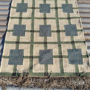Può includere: Un tappeto rettangolare con sfondo beige e un motivo a griglia di linee verdi e quadrati blu. Il tappeto ha una frangia su un'estremità. Il tappeto è realizzato in un materiale tessuto.