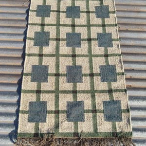 Può includere: Un runner da tavola rettangolare intrecciato con un motivo a griglia di quadrati beige chiaro accentuati da rettangoli verde acqua scuro. Il design include linee verdi che si intersecano con piccoli quadrati marrone scuro ad ogni incrocio.