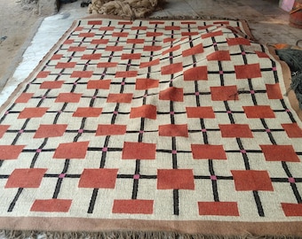 Könnte beinhalten: Ein rechteckiger Teppich mit geometrischem Muster. Der Teppich hat ein sich wiederholendes Design aus rostfarbenen Rechtecken, schwarzen Linien und kleinen rosa Quadraten auf beigem Hintergrund. Der Teppich hat einen braunen Rand und Fransen.