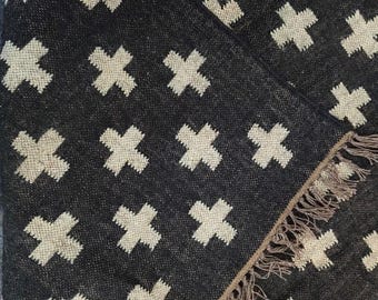 May include: A dark gray rug with a repeating pattern of off-white crosses. The rug has a fringed edge in a neutral color. The texture of the rug appears to be woven.