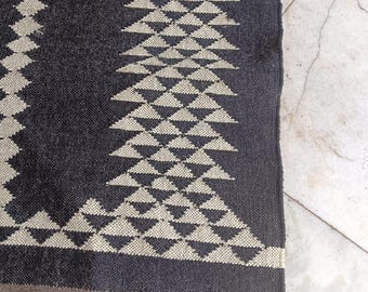May include: A dark gray woven rug with a geometric pattern of cream-colored triangles. The rug has a green and brown border with fringe along the bottom edge. The rug is made of a thick, durable material.
