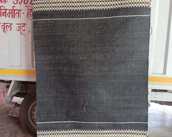 May include: A rectangular, dark gray rug with a woven texture. The rug features two bands of off-white geometric patterns and a fringe along the bottom edge. The rug is made of a natural fiber.