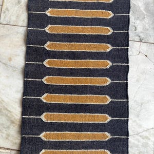 Puede incluir: Una alfombra rectangular tejida de color azul oscuro con un patrón repetido de rectángulos horizontales de color dorado. Los rectángulos están delineados en blanco y están espaciados uniformemente. La alfombra tiene un borde con flecos en la parte superior.
