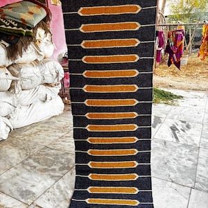 May include: A rectangular, dark gray rug with a repeating pattern of horizontal, gold-colored shapes outlined in white. The rug has a fringed edge and is being held up, showcasing its design and texture.