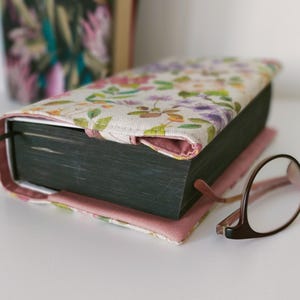 Könnte beinhalten: Ein Buch in einem Stoffumschlag mit Blumenmuster in Rosa, Violett und Grün auf cremefarbenem Grund. Das Buch hat dunkel gefärbte Seitenkanten und ein schwarzes Lesebändchen, daneben liegt eine Brille mit braunem Rahmen auf der Oberfläche.