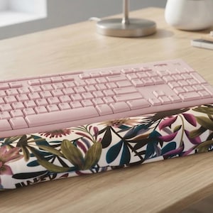 Può includere: Una tastiera per computer rosa chiaro e un mouse wireless rosa chiaro abbinato sono posizionati dietro un poggiapolsi rettangolare con una stampa botanica di foglie verde scuro e fiori rosa su sfondo bianco.