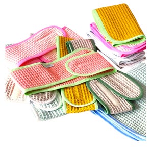 Op de afbeelding: Een collectie hoofdbanden met wafelstructuur in diverse kleuren, waaronder roze, geel, groen en blauw. Elke hoofdband is voorzien van een klittenbandsluiting en een contrasterende stoffen rand. De set bevat meerdere gevouwen en gestapelde stukken.