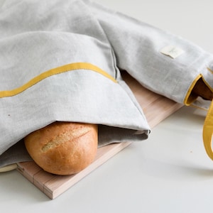 Könnte beinhalten: Eine hellgraue Brotbeutel aus Leinen mit gelbem Besatz und Griff, der ein rundes Brot enthält. Die Tasche liegt auf einem Holzschneidebrett. Die Tasche ist so konzipiert, dass sie Brot frisch hält.