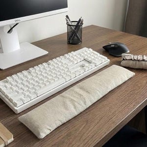 Könnte beinhalten: Eine rechteckige Handgelenkauflage aus beigem Stoff, die vor einer weißen mechanischen Tastatur liegt. Die Tastatur hat weiße Tasten und befindet sich auf einem Holztisch neben einer schwarzen Computermaus und einem kleinen, passenden beigen Stoffaccessoire.