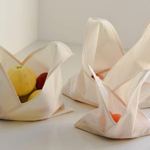 Può includere: Tre borse in tessuto color bianco sporco con un'apertura superiore triangolare e ripiegata. Le borse sono realizzate in un tessuto di colore chiaro e contengono vari frutti freschi, tra cui una mela gialla e piccoli agrumi arancioni.