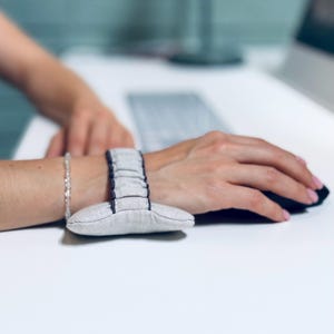Peut inclure: Un repose-poignet en tissu gris clair doté d'une base rembourrée texturée et d'une sangle élastique flexible qui se fixe autour du poignet. L'accessoire est conçu avec des coutures gris foncé visibles le long des bords des sections rembourrées.
