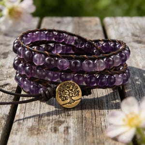 Könnte beinhalten: Wickelarmband mit Reihen aus lila Amethystperlen und dunkelbraunem Leder. Ein goldfarbener Knopf mit einem Baumdesign verschließt das Armband. Das Armband wird auf einer verwitterten Holzoberfläche präsentiert.