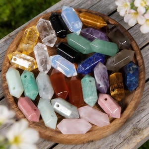 May include: A collection of polished crystal wands in various colors, including clear, yellow, black, blue, green, and pink. The crystals are arranged on a round wooden tray, showcasing their different shapes and hues. Some have holes drilled through them.