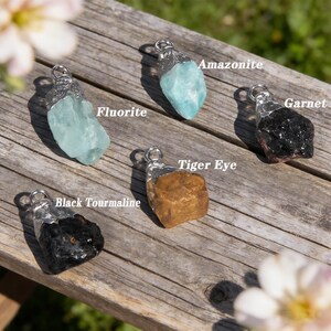 May include: Gemstone pendants featuring Fluorite, Amazonite, Garnet, Black Tourmaline, and Tiger Eye. Each stone is set in a silver-tone metal casing with a loop for a chain. The pendants are arranged on a weathered wooden surface.