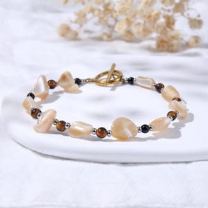 May include: A beaded bracelet featuring irregular, polished cream-colored shell fragments, round brown tiger's eye beads, and small black beads. The bracelet is finished with a gold-toned metal toggle clasp, creating a circular arrangement.