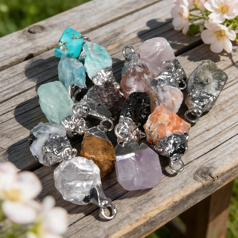 May include: A selection of raw gemstone pendants in a variety of colours, including turquoise, blue, pink, black, orange, and purple. Each stone has a silver-coloured metal cap and a loop for a chain. The pendants are displayed on a weathered wooden surface.