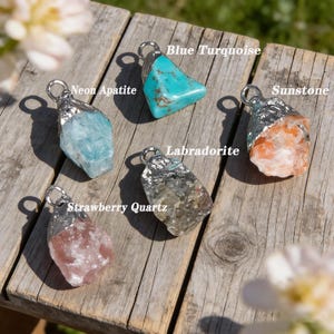 May include: Five gemstone pendants with silver-coloured metal caps are displayed on a wooden surface. The gemstones include blue turquoise, neon apatite, sunstone, labradorite, and strawberry quartz. Each stone has a unique shape and colour.