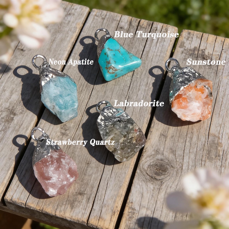 May include: Five gemstone pendants with silver-coloured metal caps are displayed on a wooden surface. The gemstones include blue turquoise, neon apatite, sunstone, labradorite, and strawberry quartz. Each stone has a unique shape and colour.