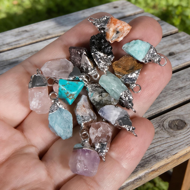 May include: A selection of gemstone pendants in diverse colours, such as pink, turquoise, and orange. Each stone has a unique shape, set with a silver-coloured metal cap and a small loop for a chain. The pendants are held in a hand.