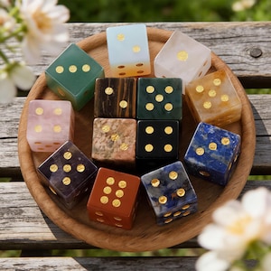 May include: A collection of twelve gemstone dice in various colors, including green, pink, and blue, with gold-colored pips. The dice are arranged on a round wooden tray, showcasing their unique textures and patterns. The dice are a standard size.