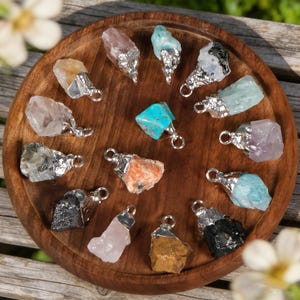 May include: A selection of gemstone pendants in diverse colours, such as turquoise, pink, and yellow, set in silver-coloured metal. The pendants are arranged on a round, wooden tray. The gemstones have a natural, rough-cut appearance.