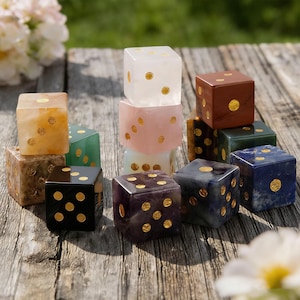May include: A collection of colorful dice made from various materials, including clear, pink, and brown stones. Each die features gold-colored dots. The dice are arranged on a weathered wooden surface, with a blurred green background.