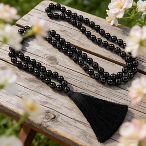 May include: A black beaded necklace with a long black tassel, resting on a weathered wooden surface. The necklace is composed of many small, round beads. The background includes soft focus white and pink flowers.