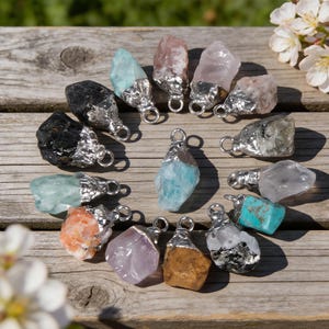 May include: A selection of gemstone pendants in diverse colours, such as black, pink, blue, and orange. Each stone is capped with a silver-coloured metal and has a loop for a chain. The pendants are displayed on a weathered wooden surface.