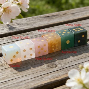 May include: A row of six gemstone dice in various colors, including clear quartz, rose quartz, and moss agate. Each die has gold-colored dots and is displayed on a wooden surface. The dice are labeled with their respective stone names.