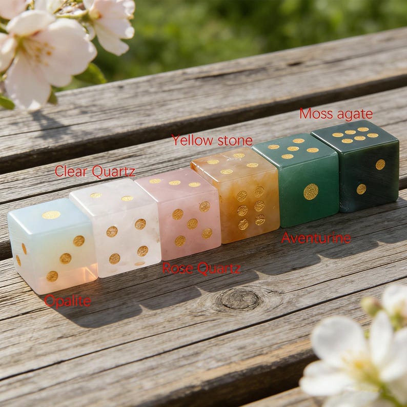 May include: A row of six gemstone dice in various colors, including clear quartz, rose quartz, and moss agate. Each die has gold-colored dots and is displayed on a wooden surface. The dice are labeled with their respective stone names.