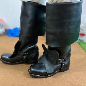 以下が含まれることがあります： 黒い革製のニーハイブーツ。つま先は四角く、装飾的なストラップが付いています。ブーツには折り返しのカフスがあり、コスチュームや歴史的再現アンサンブルの一部である可能性があります。