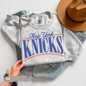 May include: A light gray crewneck sweatshirt featuring the text New York KNICKS in blue lettering, accented by orange horizontal lines above and below the team name. The garment is displayed alongside blue denim fabric and a brown felt hat.