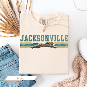 May include: An ivory short-sleeve t-shirt featuring the text JACKSONVILLE in distressed teal block letters above a graphic of a leaping black panther. The design includes horizontal teal and gold stripes. The shirt has a Comfort Colors brand label.