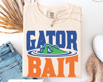 May include: An ivory short-sleeve t-shirt featuring a graphic print with the blue text GATOR above a green alligator head emerging from blue water ripples, and the orange text BAIT below. The shirt has a Comfort Colors brand neck label.