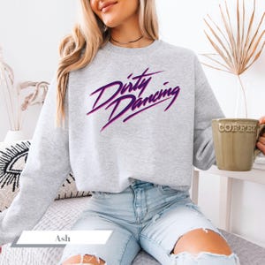 Peut inclure: Sweat-shirt gris clair avec l'inscription "Dirty Dancing" en script violet et rose. Le sweat-shirt à col rond est porté avec un jean délavé clair. Une tasse verte avec le mot "Coffee" est tenue à la main.