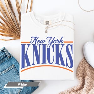 May include: White t-shirt with the text "New York KNICKS" in blue and orange. The shirt has a crew neck and short sleeves. The shirt is made of a soft, comfortable material.